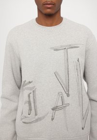 Ljusgrå sweatshirt med en texturerad, sydd design och abstrakt text i mörkgrått. Ribbad halsringning och ärmslut. Mjuk tyg.