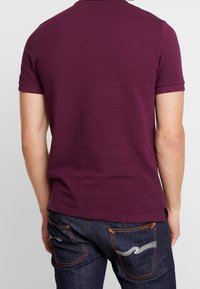 Burgundská polo košile s krátkým rukávem z měkké bavlny, s jednoduchým designem. V kombinaci s tmavými džíny s oranžovými švy.