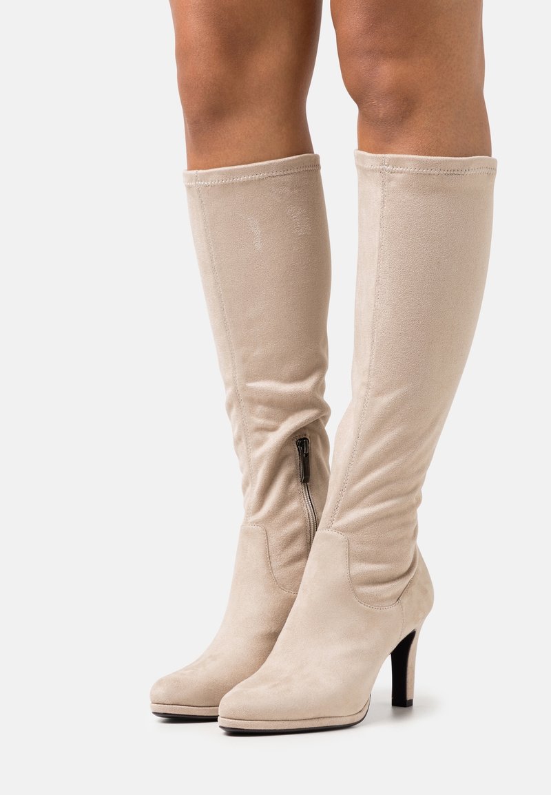 Stivali alti beige in suede con zip laterale e tacchi alti indossati su gambe scoperte su sfondo bianco.