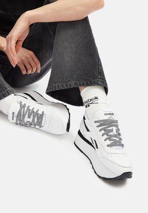 Baltos Reebok kedai su sportu su pilkais akcentais, tekstūruotomis raištelėmis ir masyviu padu. Juodi akcentai kulno srityje ir perforuota pirštų dalis, skirta oro pralaidumui.