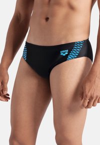 Bañador tipo slip negro con un patrón geométrico azul en el lado izquierdo y el logo "arena" en azul en la cinturilla. Textura lisa, diseño ajustado.