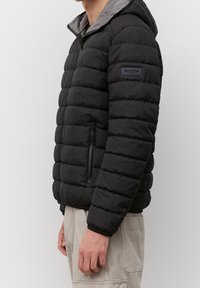 Schwarze Puffjacke mit Kapuze, mit gesteppten Abschnitten, einer Reißverschlusstasche an der Seite und einem grauen Innenfutter. Markenlabel am Arm.
