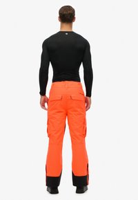 Man in felroze cargobroek en een strakke zwarte lange mouwen shirt, staand met zijn rug naar de camera tegen een eenvoudige witte achtergrond.