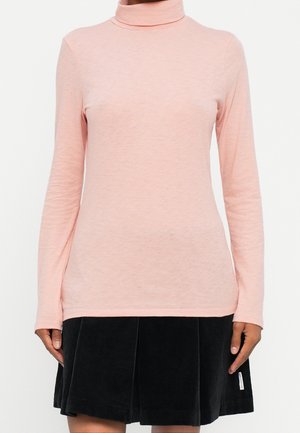 Vrouw met een lichtroze coltrui met lange mouwen gecombineerd met een zwarte geplooide rok, staand tegen een witte achtergrond.