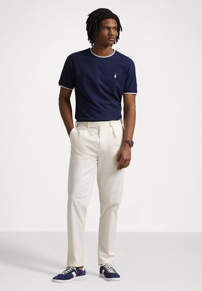 T-shirt azul de gola redonda com detalhes brancos, calças brancas plissadas e ténis azuis com acentos vermelhos e brancos. Conjunto casual adequado para a primavera.