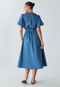 Denim maksi obleka v srednje modri barvi, kratki naborki na rokavih, elastičen pas, detajl v obliki ključavnice na hrbtu in svilnato krilo. Gladka tekstura, sproščen dizajn.