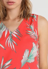 Top rojo sin mangas con escote en V, que presenta patrones de hojas tropicales en verde y rosa sobre una tela suave. Los detalles incluyen ornamentación en los hombros.
