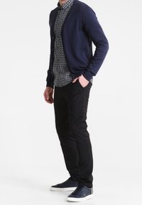 Marinblå cardigan med knappframsida, bar över en svart och vit rutad skjorta, ihop med svarta byxor och marinblå sneakers. Enkel, modern design.