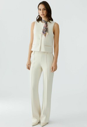 Vrouw die een mouwloos crèmekleurig vest en bijpassende hoge taillebroek draagt, met een kleurrijke sjaal om haar nek, staand tegen een effen achtergrond.