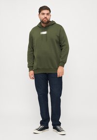 Sudadera con capucha de color verde oliva hecha de tela suave, con un parche de logo en el frente y cordones ajustables, combinada con jeans de color azul oscuro y zapatillas negras.