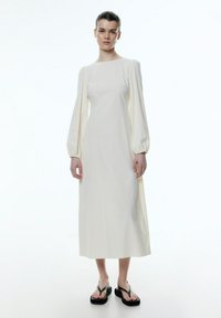 Cremfarbendes Midi-Kleid mit langen Puffärmeln und quadratischem Ausschnitt. Hergestellt aus leichtem Stoff. Kombiniert mit schwarzen Plateausandalen.