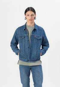 Veste en denim avec un motif, fermeture à boutons sur le devant, deux poches poitrine et ourlet côtelé. Portée sur un t-shirt gris et accompagnée de jeans.