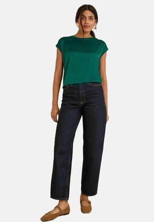 Türkiser Satin-Top mit kurzen Ärmeln, kombiniert mit hoch taillierten dunklen Jeans. Das Model trägt beige Ballerinas und schlichte Ohrringe.