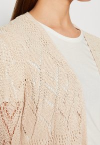 Gros plan d'une personne portant un cardigan beige en maille ouverte sur un t-shirt blanc uni à col rond, montrant des motifs de tricot détaillés et la texture.