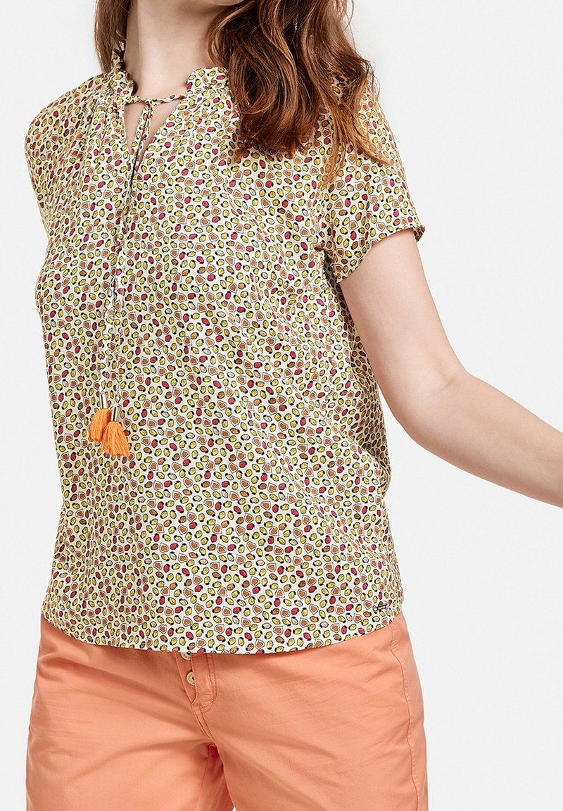 Femme portant un chemisier à manches courtes et à motifs avec un lien et des pompons, associé à un pantalon couleur saumon sur fond uni.
