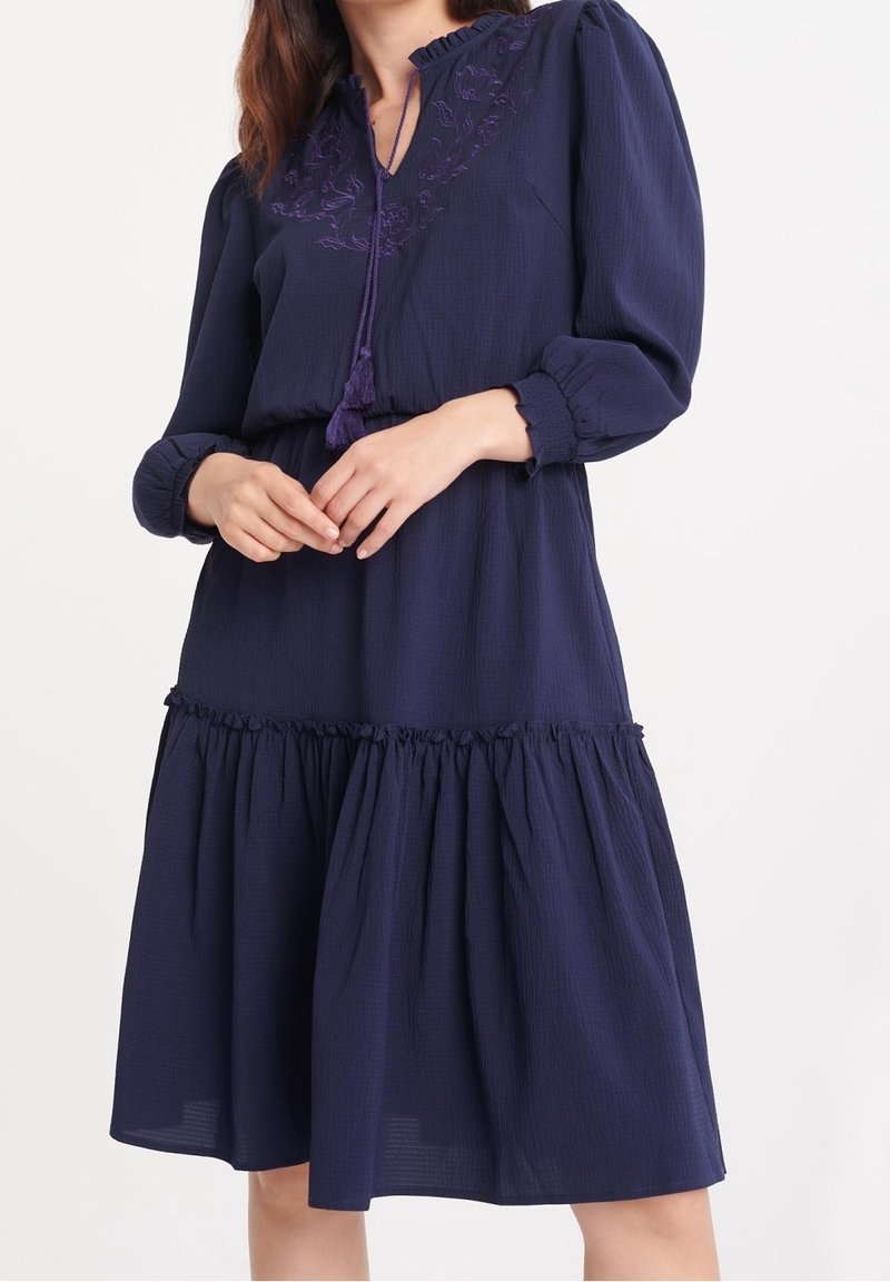 Femme portant une robe bleu marine à manches longues avec jupe froncée et encolure brodée, mains doucement jointes à la taille.