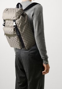 Sac à dos en tissu beige avec un motif, doté de bretelles noires ajustables et d'une poche zippée à l'avant. Porté sur un pull gris et un pantalon rayé.