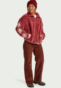 Sudadera de forro polar en burdeos con media cremallera, bolsillos frontales y patrones florales blancos en las mangas; combinada con pantalones de pana color óxido.