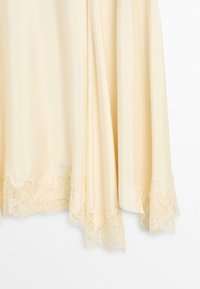 Crèmekleurige zijden stof met kanten rand langs de onderkant, zacht gedrapeerd tegen een effen witte achtergrond.