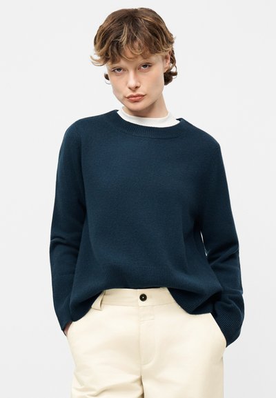 Jeune personne aux cheveux courts et bouclés portant un pull bleu foncé sur une chemise blanche et un pantalon couleur crème, mains dans les poches.