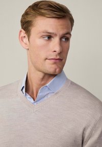 Svijetlo bež v-neck pulover od mekanog pletiva, složen preko plave košulje s ovratnikom, s glatkom teksturom i jednostavnim dizajnom.
