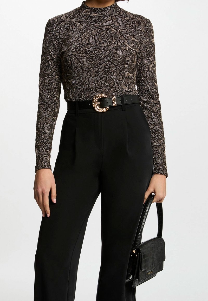Femme portant un haut à manches longues à motif floral métallique, un pantalon noir taille haute avec une ceinture à boucle dorée, tenant un sac à main noir.