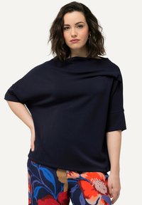 Top drappe blu navy con maniche corte, caratterizzato da una vestibilità ampia e una consistenza morbida, abbinato a pantaloni a fantasia floreale colorati.