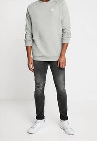 Ljusgrå sweatshirt med rund halsringning och en liten vit logotyp, tillsammans med mörkgrå slitna jeans och vita sneakers.