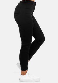 Mujer vestida con jeans ajustados de cintura alta negros y zapatillas blancas de perfil bajo, de pie con una pierna doblada contra un fondo liso.