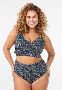 Hymyilevä nainen, jolla on kiharat hiukset ja päällään tummansininen kukkakuvioinen kahden osan uimapuku, jossa röyhelöt, seisoo käsivarret lanteilla valkoista taustaa vasten.