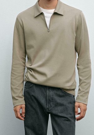 Uomo che indossa un pullover beige a manica lunga con mezza zip sopra una camicia bianca e jeans grigio scuro, in piedi davanti a uno sfondo grigio chiaro.