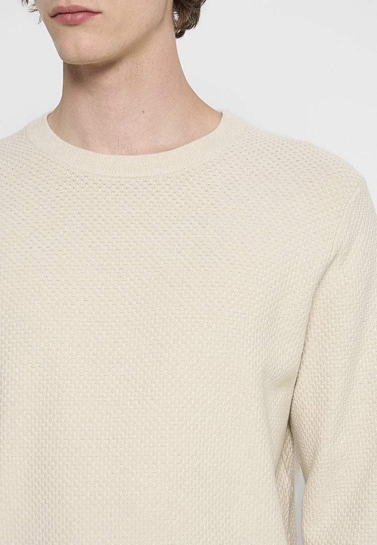 Ljusbeige texturerad tröja med rund halsringning. Stickmönstret har en subtil upphöjd design, vilket ger en mjuk och andningsbar känsla.