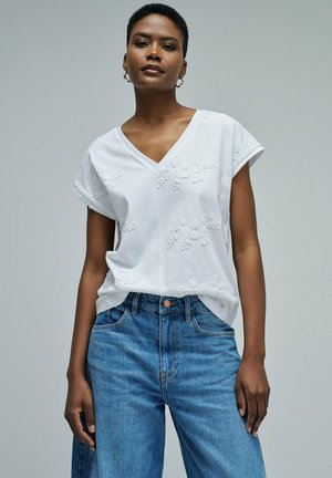 Mujer negra con blusa blanca de manga corta con escote en V y bordados, y vaqueros azules de cintura alta, de pie frente a un fondo liso.