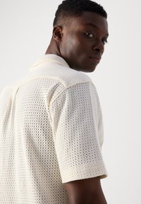 Polo a maglia in crema con un design a rete aperta e texture. Presenta un colletto morbido e maniche corte, offrendo un comfort traspirante.