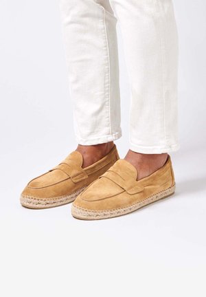 Person iført lyse beige ruskindsluffer med vævede espadrille-såler og hvide bukser mod en ensfarvet baggrund.