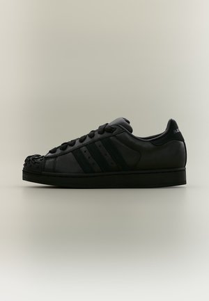 Μαύρο χαμηλό sneaker με κορδόνια, αστέρια ως ανοίγματα στις πλευρικές ρίγες και υφασμένη λαστιχένια μύτη, σε απλό ανοιχτό φόντο.