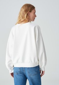 Felpa bianca corta con scollatura tonda, spalle scese e polsini a costine, abbinata a jeans di denim blu. Texture liscia, design semplice.