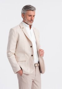 Blazer de lino beige claro con dos botones y bolsillos con solapa, llevado sobre una camisa blanca, combinado con pantalones beige claro y un cinturón estampado.