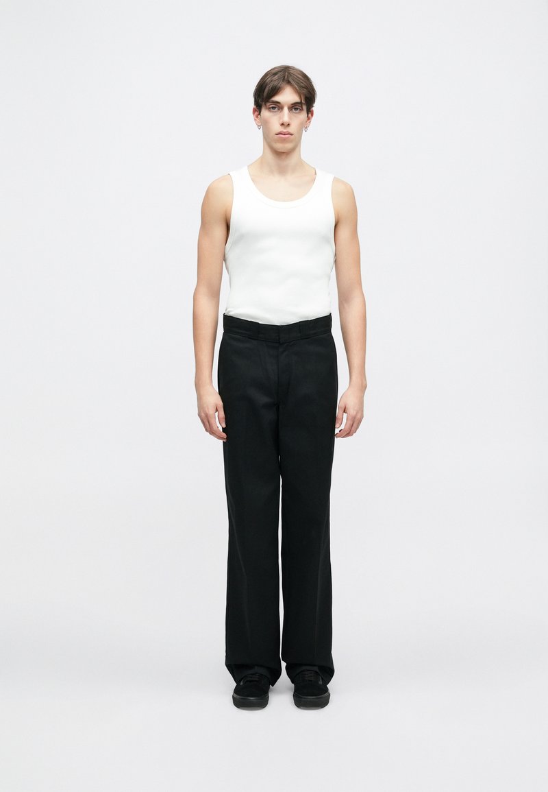 Hvit ermeløs tanktopp kombinert med svarte, vide bukser. Modellen står rett mot en ensfarget hvit bakgrunn. Svarte sko fullfører antrekket.