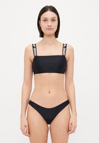 Black bikini with a square-cut top and thin straps featuring the brand "arena" in white. The bottom has a high-cut design, smooth texture.