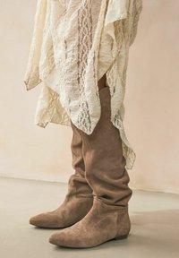 Botas de ante marrón hasta la rodilla con puntera afilada, presentando una textura suave y un ligero detalle de caída, combinadas con una chal blanca texturizada.