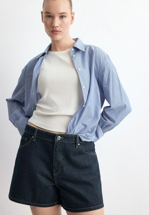 Kobieta ubrana w biały bezrękawnik, niebiesko-białą koszulę w paski na guziki oraz ciemne szorty dżinsowe na jednolitym białym tle.