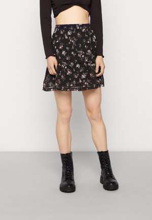Femme portant une jupe courte noire à fleurs, un crop top noir et des bottes de combat noires à lacets, posant devant un fond uni.