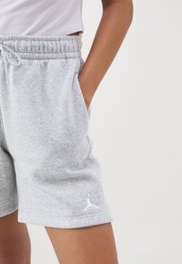 Personne portant un short gris clair à cordon de serrage avec le logo Jordan sur la jambe gauche, la main posée de manière décontractée dans la poche droite, sur un fond uni.