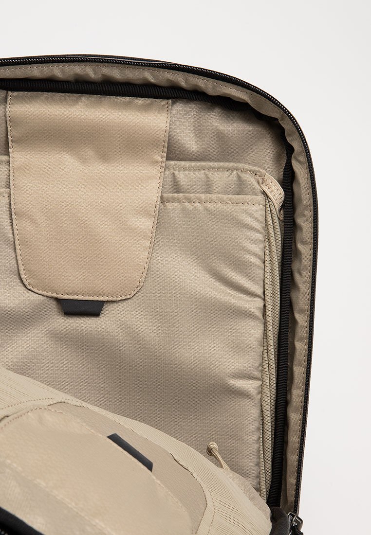 Comparto per laptop imbottito beige all'interno di uno zaino, con un flap di chiusura e una cerniera nera visibile lungo il bordo.