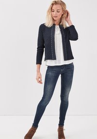 Vadderad marinblå jacka med rund halsringning och framdragkedja, bärs över en vit topp, ihop med blå slim fit-jeans och bruna ankelskor.