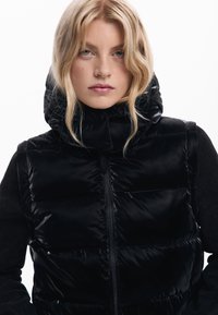 Gilet imbottito nero con collo alto e chiusura con cerniera; tessuto lucido; sezioni imbottite; abbinato a una maglia nera a maniche lunghe.