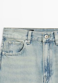 Jean en denim bleu clair au niveau de la taille, montrant le bouton, les passants de ceinture, les poches avant et la poche monnaie avec des détails de coutures visibles.