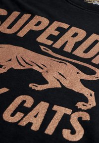 Camiseta de algodón negra con estampado de purpurina metálica color rosa dorado de la silueta de un gato y el texto "SUPERDRY CATS". Diseño audaz y texturizado.