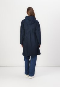 Parka impermeabile blu navy con cappuccio, vita stretta, maniche lunghe e orlo dritto, abbinata a jeans a zampa d'elefante e sneaker bianche.